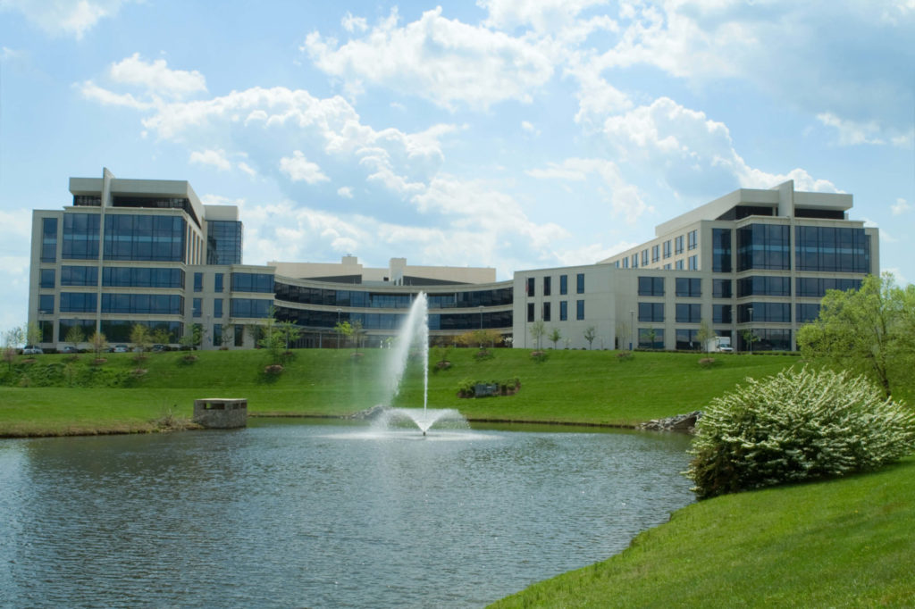 Corporate Campus with office buildings with a grass, trees, a pond and fountain in front of the building with blue sky in background