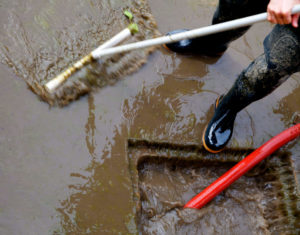 Upshot image of worker in rubber boots using a tool, cleaning after a flood, by moving dirty water and debris into a drainage system with pump and hose