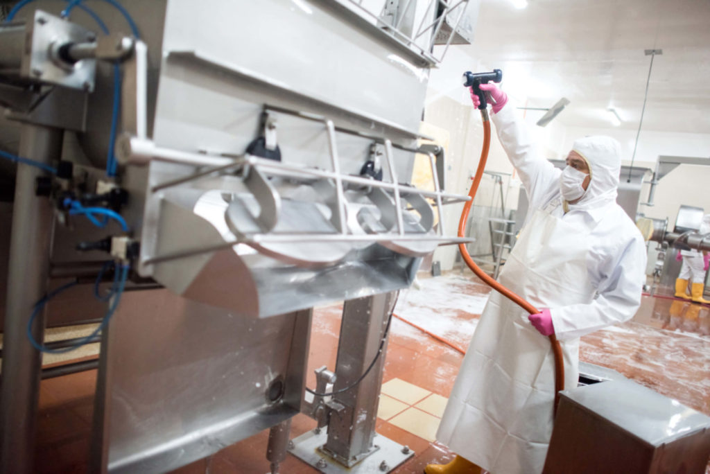 Man in food processing plant wearing coveralls, apron, mask, hood and rubber boots, holding a hose washing down a machine