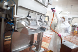 Man in food processing plant wearing coveralls, apron, mask, hood and rubber boots, holding a hose washing down a machine