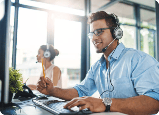 Cropped shot of a smiling young man working at computer wearing a headset with a female colleague in the background