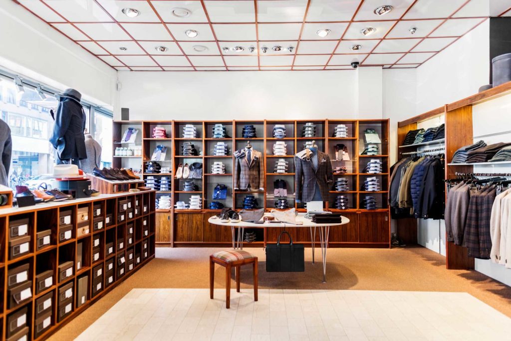 Interior view of high-end men’s clothing store with suits, dress shirts, belts, shoes, fedora style hats, jackets with mannequins dressed in suits.
